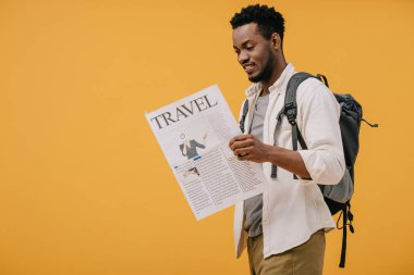 cheerful african american man standing with backpack and reading travel newspaper isolated on orange clipart