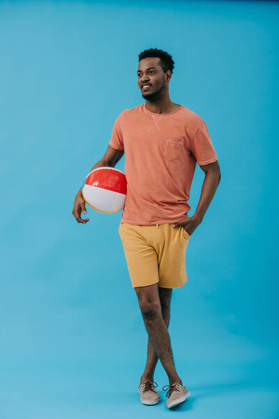 счастливый африканский американец, стоящий с рукой в кармане и держащий пляжный мяч на голубом
 