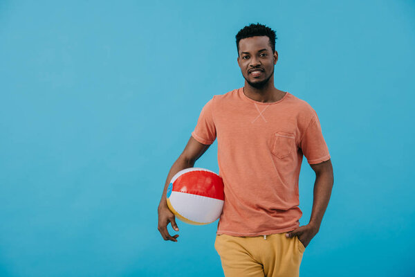 счастливый африканский американец, стоящий с рукой в кармане и держащий пляжный мяч изолирован на голубом
 