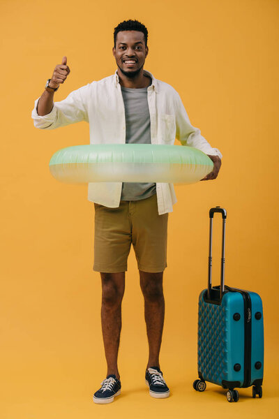 cheerful african american man standing with swim ring and luggage while showing thumb up on orange