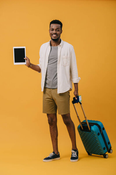cheerful african american man standing with luggage and holding digital tablet with blank screen on orange