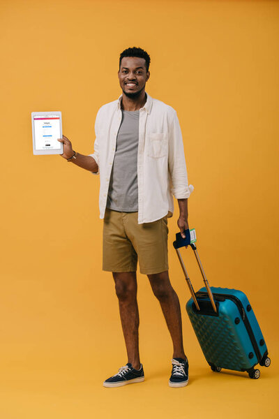 KYIV, UKRAINE - JUNE 27, 2019: cheerful african american man standing with luggage and holding digital tablet with instagram app on screen on orange