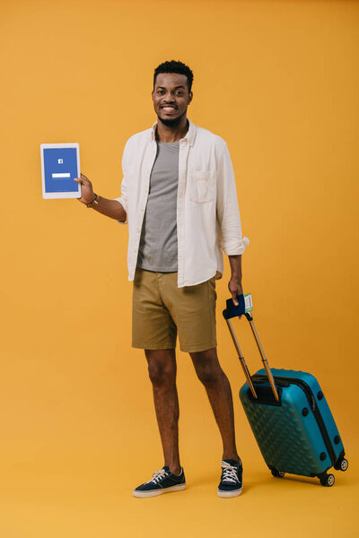 KYIV, UKRAINE - JUNE 27, 2019: cheerful african american man standing with luggage and holding digital tablet with facebook app on screen on orange