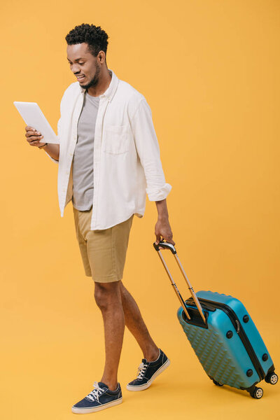 cheerful african american man holding digital tablet and walking with luggage on orange