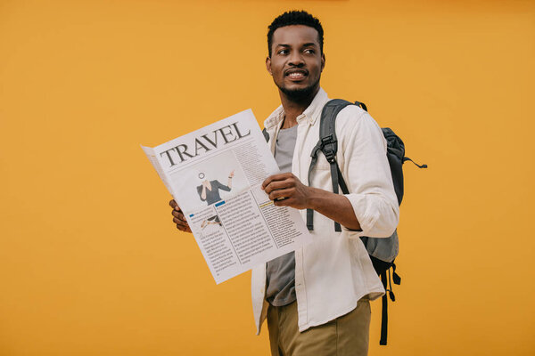 cheerful african american man standing with backpack and holding travel newspaper isolated on orange