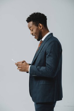 gri izole akıllı telefon kullanarak takım elbiseli sürpriz Afro-Amerikan adam yan görünümü 