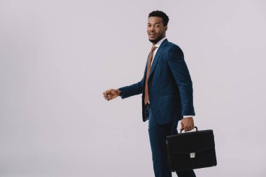 happy african american man in suit holding briefcase isolated on grey  clipart
