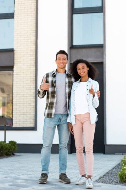 full length view of african american husband and wife showing thumbs up while standing near new house and looking at camera clipart