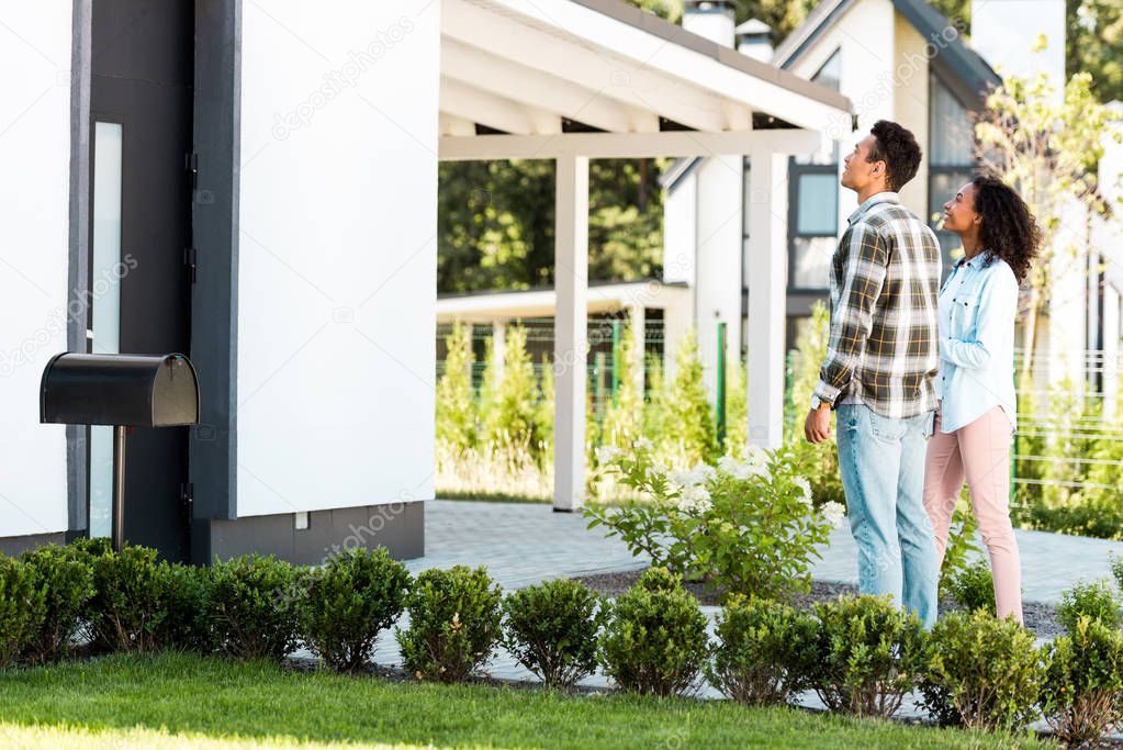 full length view of african american man and woman looking at house while standing near lawn