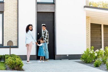 Afro-Amerikan baba çocuk ve anne kameraya bakarak el ele tutuşurken yeni evin yanında duran ailenin tam uzunlukta görünümü