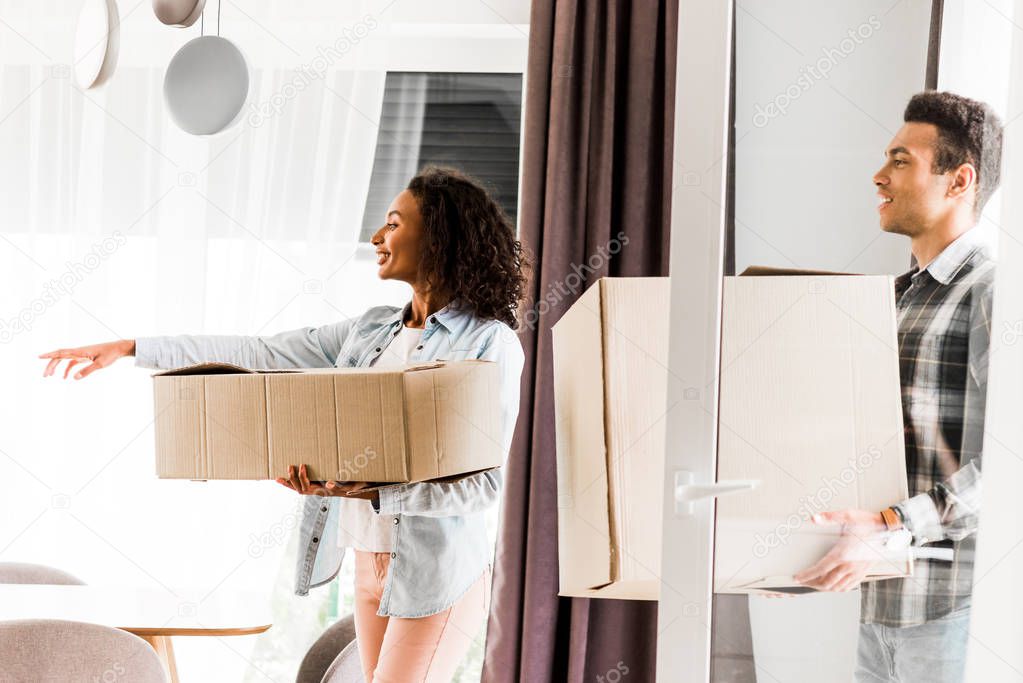 african american husband and wife walking into house and holding boxes while woman pointing with hand