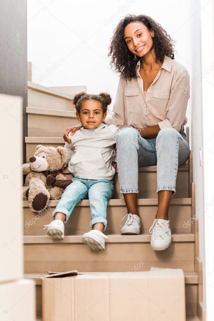 full length view of mother and kid sitting at stairs, smiling and looking at camera