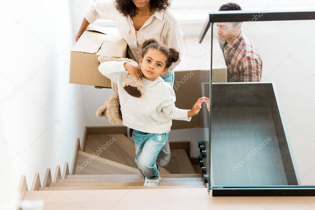 cropped view of african american woman going upstairs with box while daughter holding toy and father walking after mother 