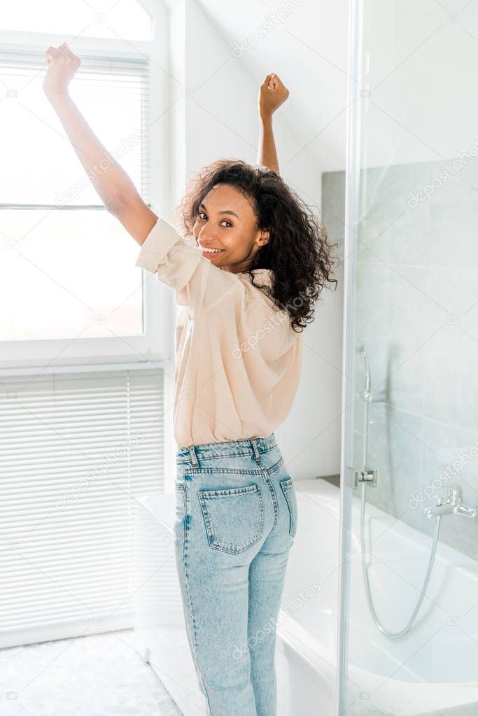 beautiful african american woman looking at camera while standing in bathroom with hands in air