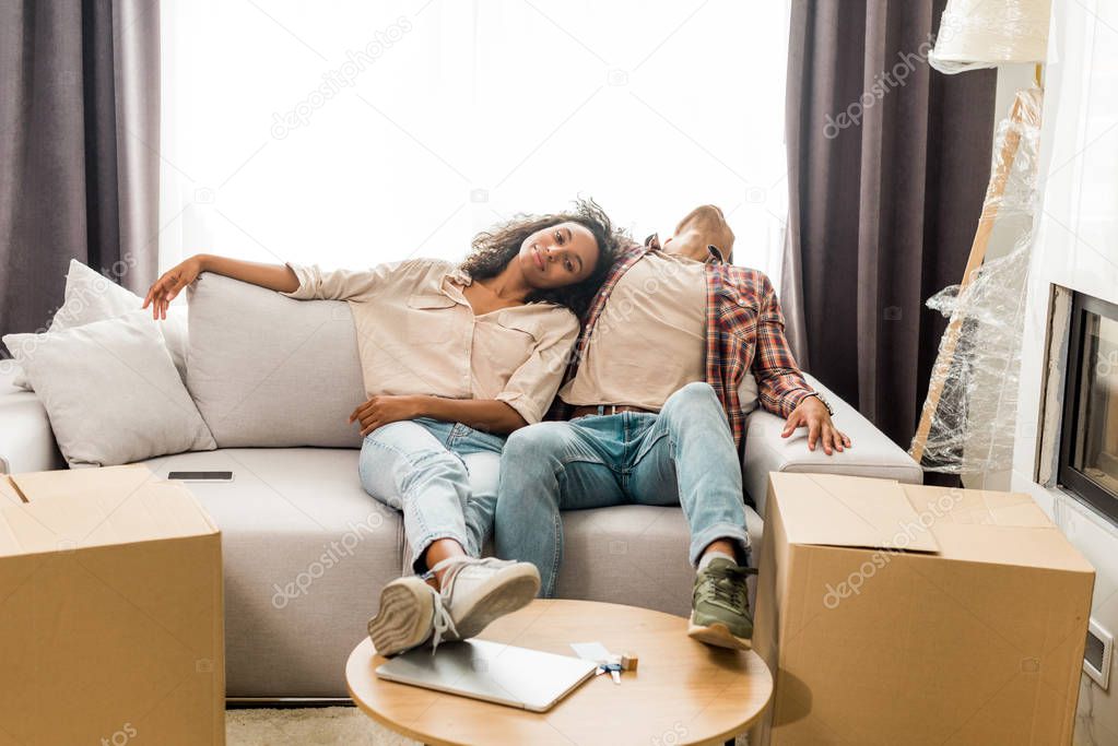 full length view of husband sleeping on sofa while wife sitting near man and looking at camera 
