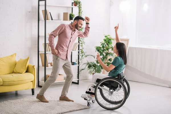 happy disabled woman on wheelchair dancing with cheerful boyfriend at home