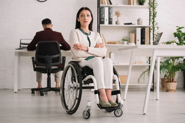 自信を持って車椅子に座っている自信のある障害のあるビジネスウーマンは 背景に取り組んでいるコレゲの近くに腕を組んで座っています — ストック写真
