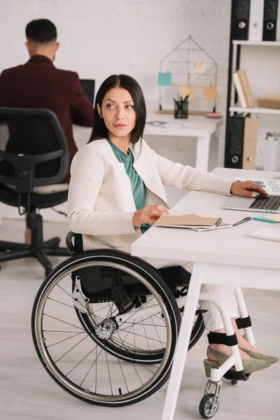魅力的な障害のあるビジネスウーマンは 背景に同僚の近くの職場で車椅子に座っている間に目をそらす — ストック写真