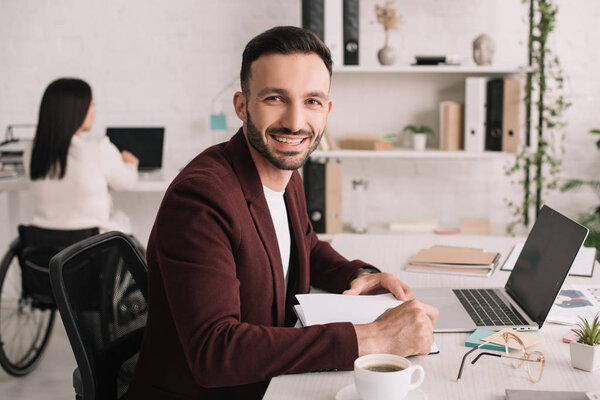 cheerful businessman looking at camera while sitting at workplace near disabled businesswoman