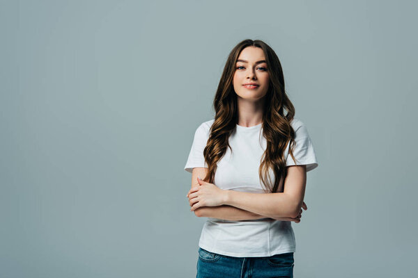 positive beautiful girl in white t-shirt with crossed arms looking at camera isolated on grey