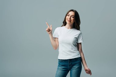 smiling beautiful girl in white t-shirt pointing with finger aside isolated on grey clipart