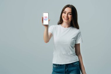 Kiev, Ukrayna - 6 Haziran 2019: beyaz tişörtlü mutlu güzel kız, gri üzerinde izole edilmiş apple müzik simgesiolan akıllı telefonu gösteriyor