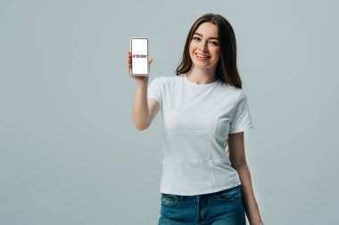 KYIV, UKRAINE - JUNE 6, 2019: happy beautiful girl in white t-shirt showing smartphone with tinder app isolated on grey clipart