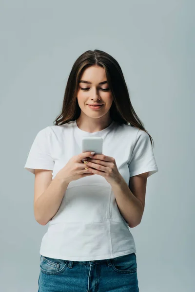 微笑美丽的女孩在白色T恤使用智能手机隔离在灰色 — 图库照片
