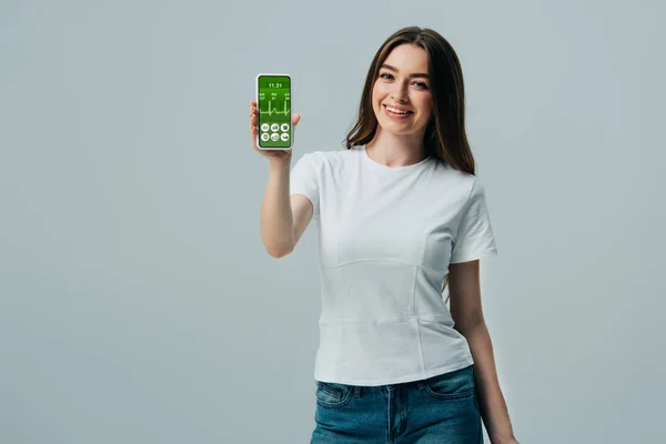 灰色で隔離されたヘルスケアアプリでスマートフォンを示す白いTシャツで幸せな美しい女の子 — ストック写真