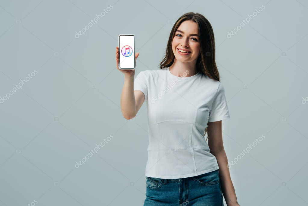 KYIV, UKRAINE - JUNE 6, 2019: happy beautiful girl in white t-shirt showing smartphone with apple music icon isolated on grey