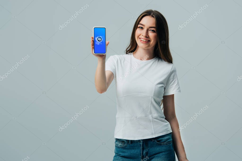 KYIV, UKRAINE - JUNE 6, 2019: happy beautiful girl in white t-shirt showing smartphone with shazam app isolated on grey