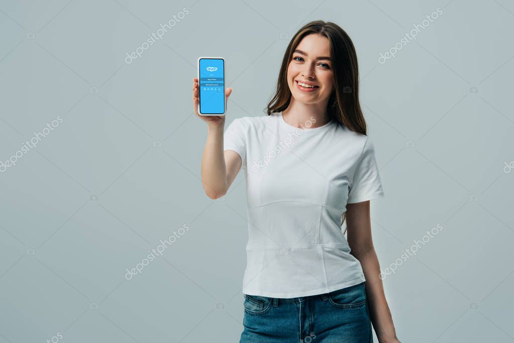 KYIV, UKRAINE - JUNE 6, 2019: happy beautiful girl in white t-shirt showing smartphone with skype app isolated on grey