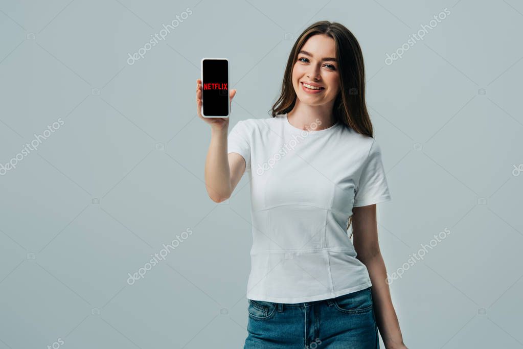 KYIV, UKRAINE - JUNE 6, 2019: happy beautiful girl in white t-shirt showing smartphone with Netflix app isolated on grey