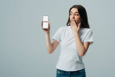 KYIV, UKRAINE - JUNE 6, 2019: shocked beautiful girl in white t-shirt showing smartphone with Tinder app isolated on grey clipart