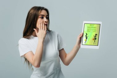 shocked beautiful girl in white t-shirt showing digital tablet with online shopping app isolated on grey clipart