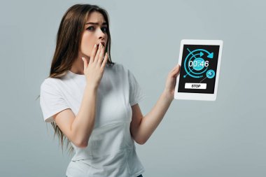 shocked beautiful girl in white t-shirt showing digital tablet with stopwatch app isolated on grey clipart