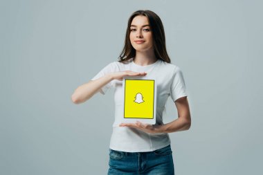 Kiev, Ukrayna - 6 Haziran 2019: gri üzerinde izole Snapchat uygulaması ile dijital tablet gösteren beyaz t-shirt gülümseyen güzel kız