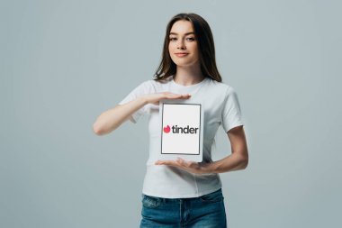 Kiev, Ukrayna - 6 Haziran 2019: gri üzerinde izole Tinder uygulaması ile dijital tablet gösteren beyaz t-shirt gülümseyen güzel kız