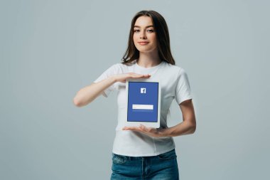 Kiev, Ukrayna - 6 Haziran 2019: gri üzerinde izole Facebook uygulaması ile dijital tablet gösteren beyaz t-shirt gülümseyen güzel kız