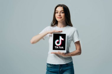Kiev, Ukrayna - 6 Haziran 2019: gri üzerinde izole Tik Tok uygulaması ile dijital tablet gösteren beyaz t-shirt gülümseyen güzel kız