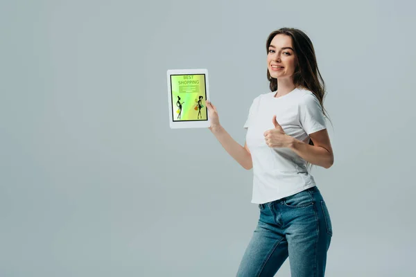 微笑美丽的女孩在白色T恤显示数字平板电脑与在线购物应用程序 并显示大拇指隔离在灰色 — 图库照片