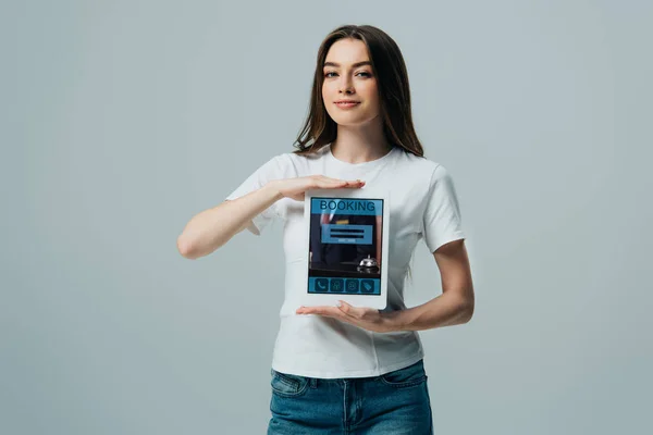 灰色で隔離された予約アプリでデジタルタブレットを示す白いTシャツで笑顔の美しい女の子 — ストック写真