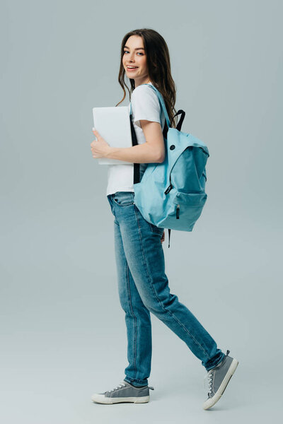 full length view of pretty smiling girl in jeans with blue backpack holding digital tablet isolated on grey