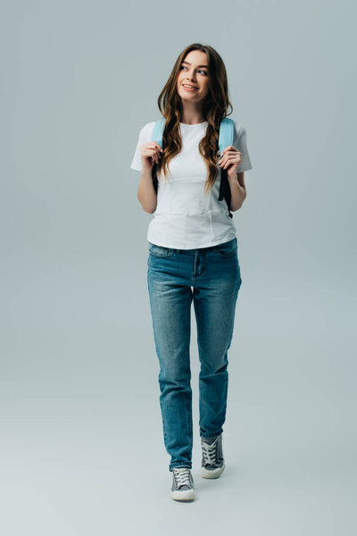 Вид в полный рост мечтательной симпатичной улыбающейся девушки с синим рюкзаком, изолированным на сером
