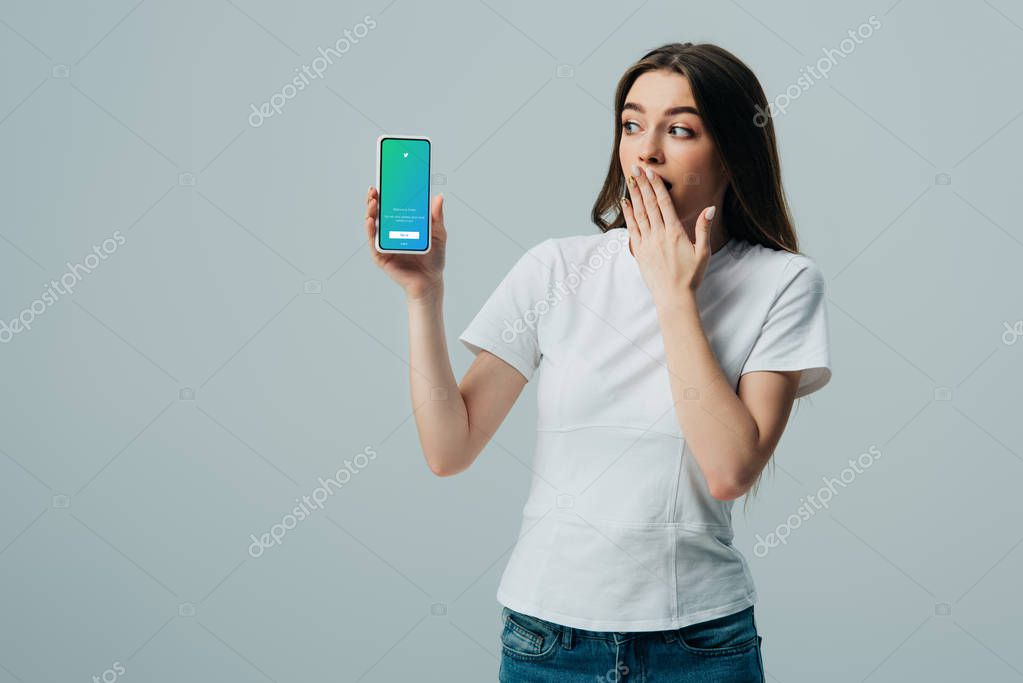 KYIV, UKRAINE - JUNE 6, 2019: shocked beautiful girl in white t-shirt showing smartphone with twitter app isolated on grey