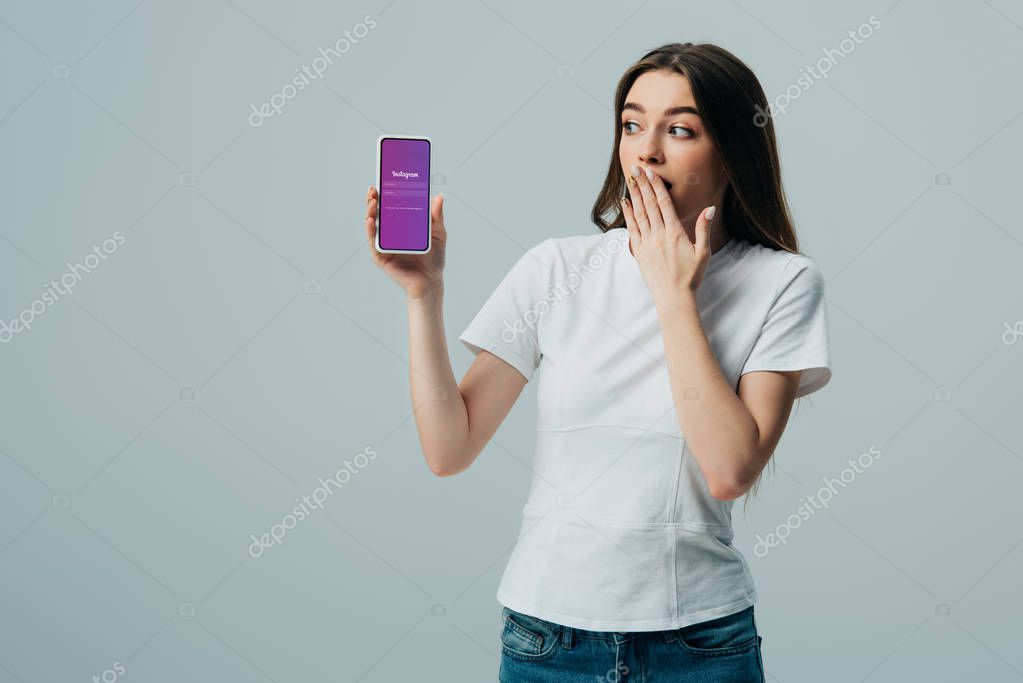 KYIV, UKRAINE - JUNE 6, 2019: shocked beautiful girl in white t-shirt showing smartphone with Instagram app isolated on grey