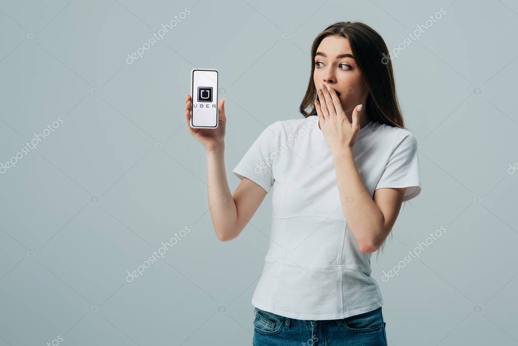KYIV, UKRAINE - JUNE 6, 2019: shocked beautiful girl in white t-shirt showing smartphone with Uber app isolated on grey