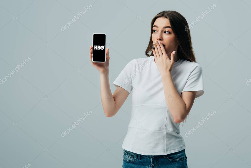 KYIV, UKRAINE - JUNE 6, 2019: shocked beautiful girl in white t-shirt showing smartphone with HBO app isolated on grey