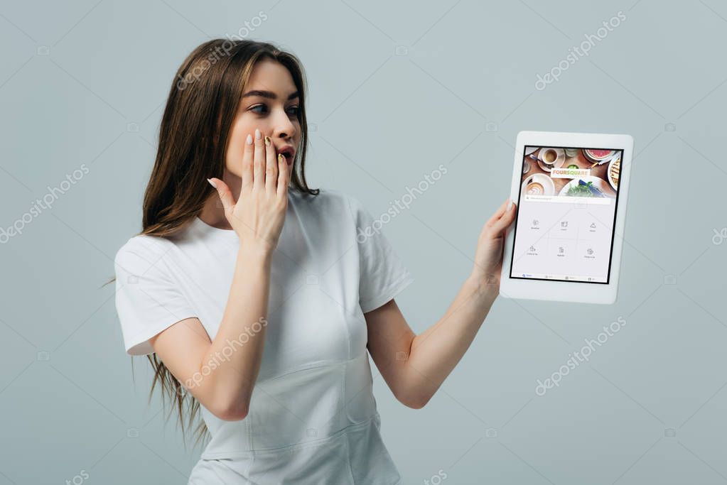KYIV, UKRAINE - JUNE 6, 2019: shocked beautiful girl in white t-shirt showing digital tablet with foursquare app isolated on grey