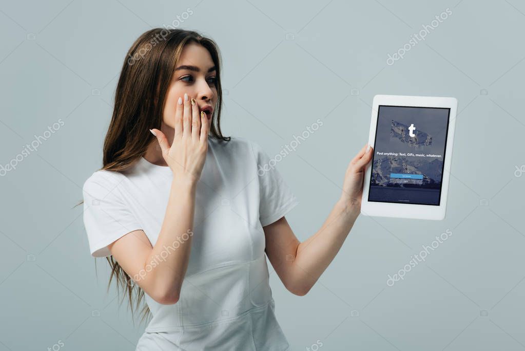 KYIV, UKRAINE - JUNE 6, 2019: shocked beautiful girl in white t-shirt showing digital tablet with Tumblr app isolated on grey